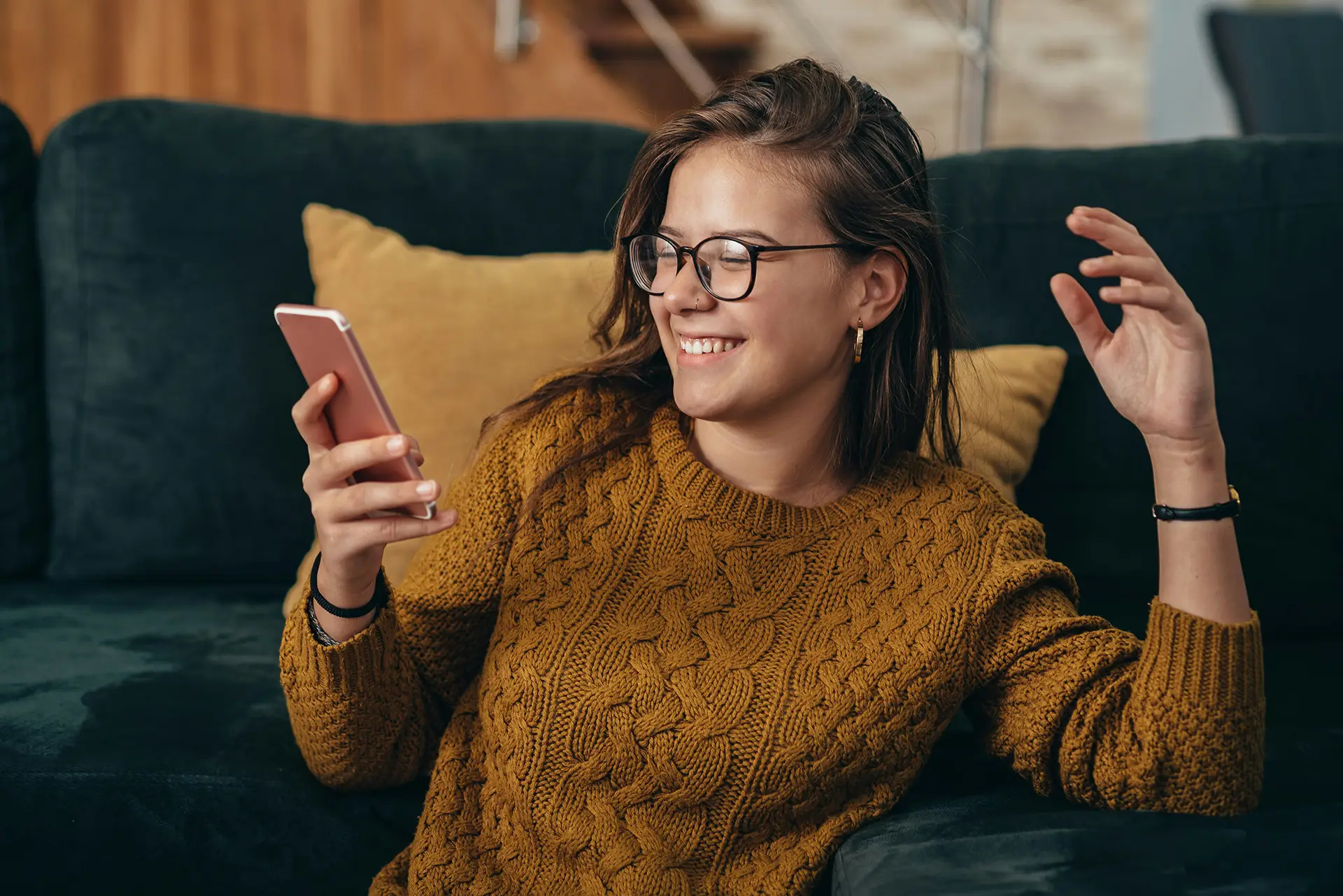 Smiling woman in glasses and brown knit sweater looking at phone while sitting on dark couch.