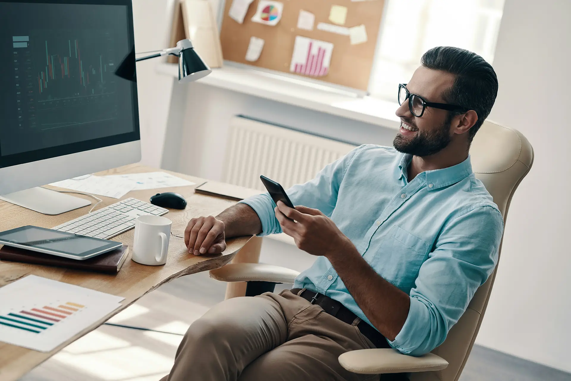 Smiling man in glasses sitting at desk, holding smartphone with computer and coffee cup on desk.