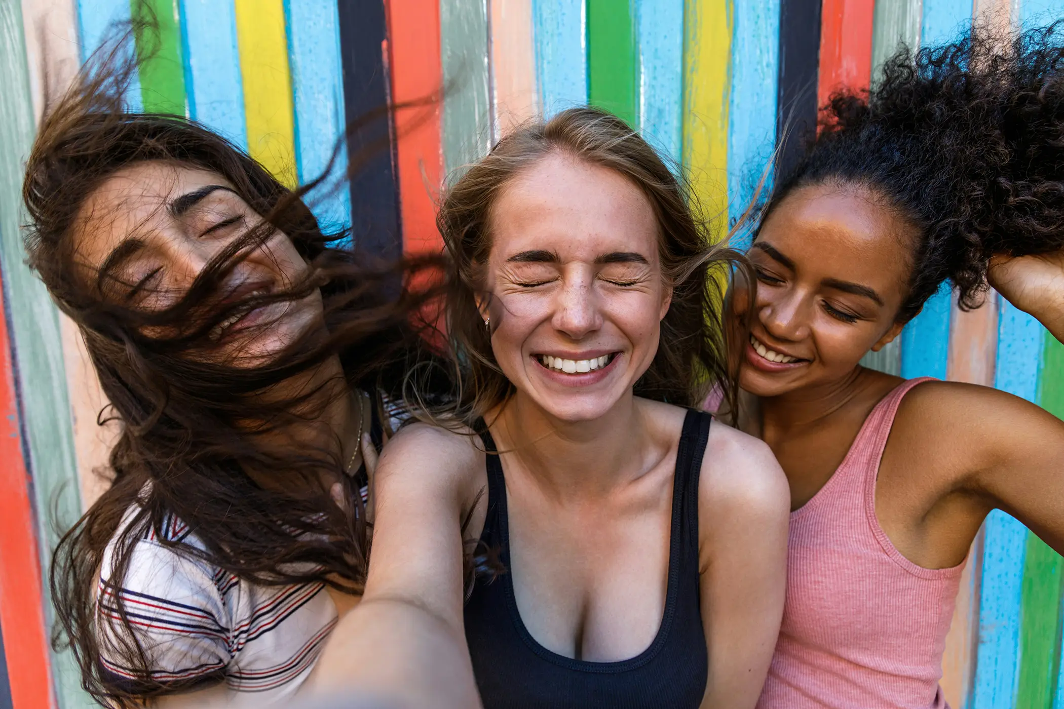 Three smiling women taking a selfie in front of a colorful striped wall with hair blowing.