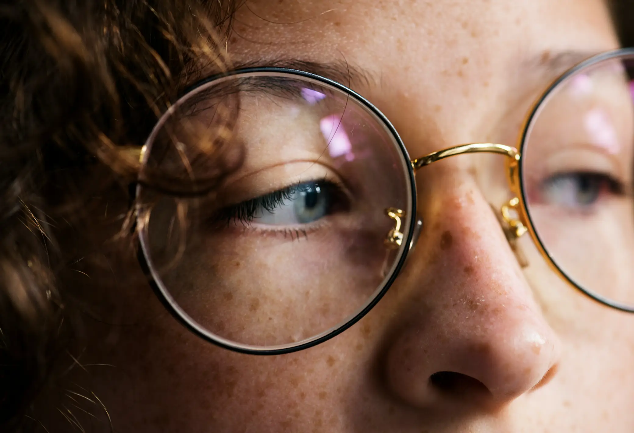 Close-up of a person's blue eyes behind round gold-rimmed glasses with reflections.